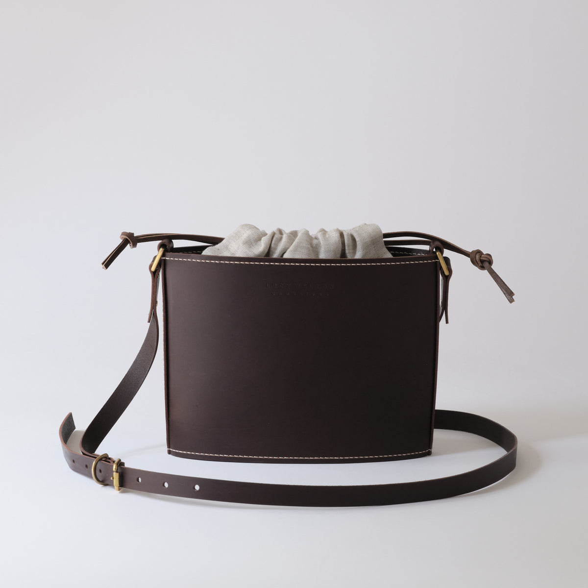 A dark brown leather bucket bag with a drawstring closure and a matching adjustable shoulder strap, shown on a clean light gray background.