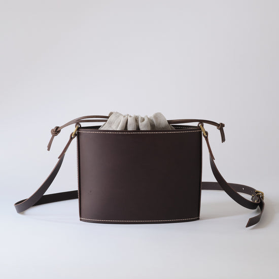 A dark brown leather bucket bag with a drawstring closure and a matching adjustable shoulder strap, shown on a clean light gray background.