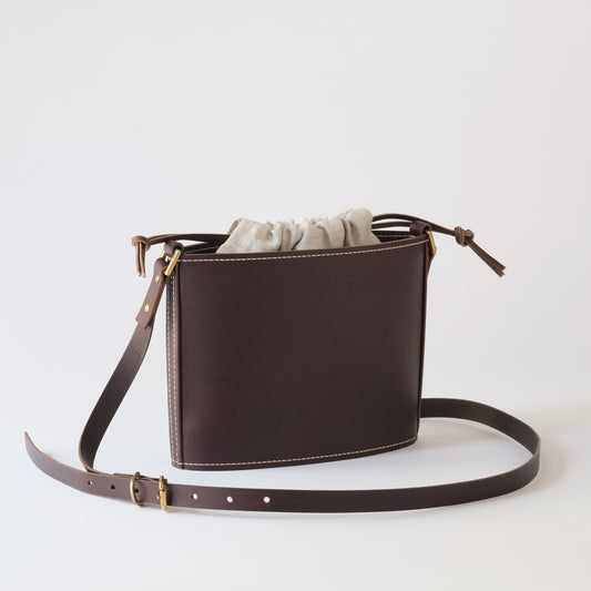 A dark brown leather bucket bag with a drawstring closure and a matching adjustable shoulder strap, shown on a clean light gray background.