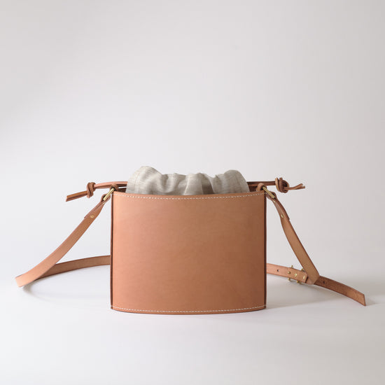 A light tan leather crossbody bag with a drawstring top and a light gray fabric pouch inside, displayed on a clean white studio background.