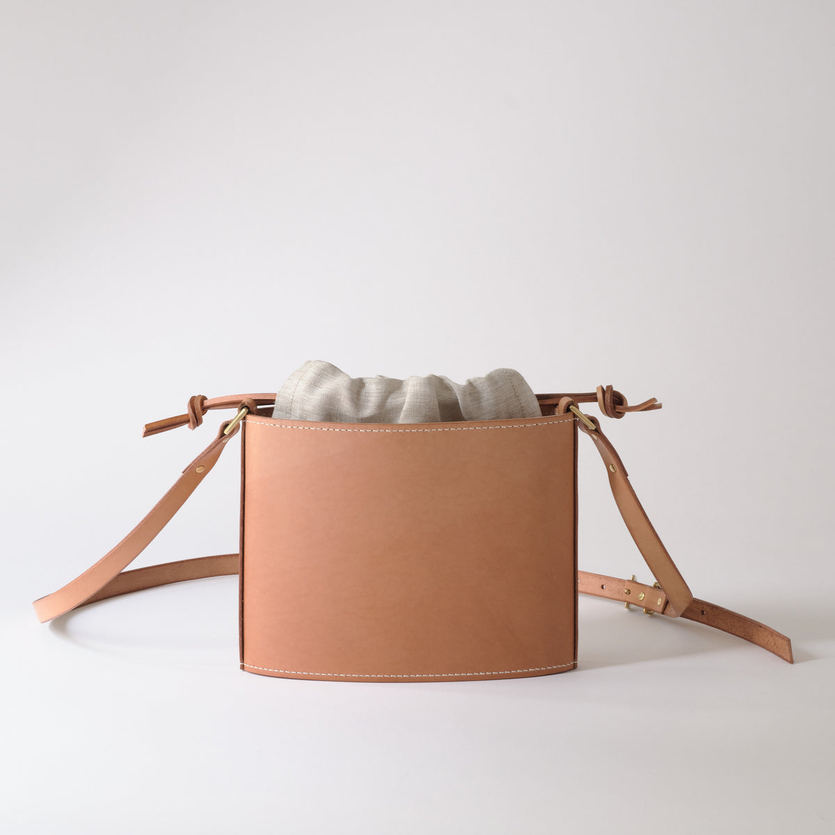 A light tan leather crossbody bag with a drawstring top and a light gray fabric pouch inside, displayed on a clean white studio background.