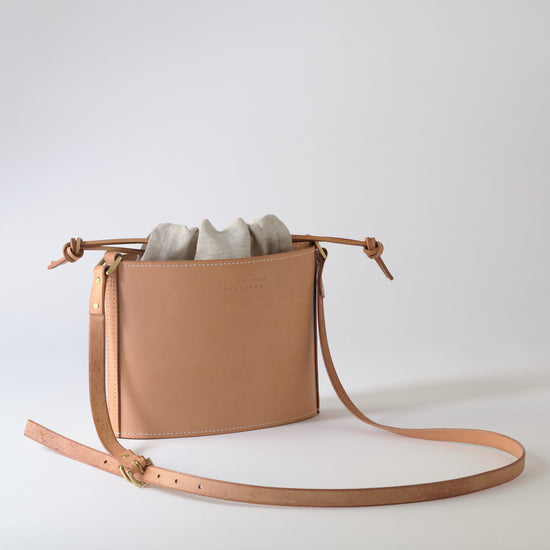 A light tan leather crossbody bag with a drawstring top and a long adjustable strap, shown on a clean light gray studio background.