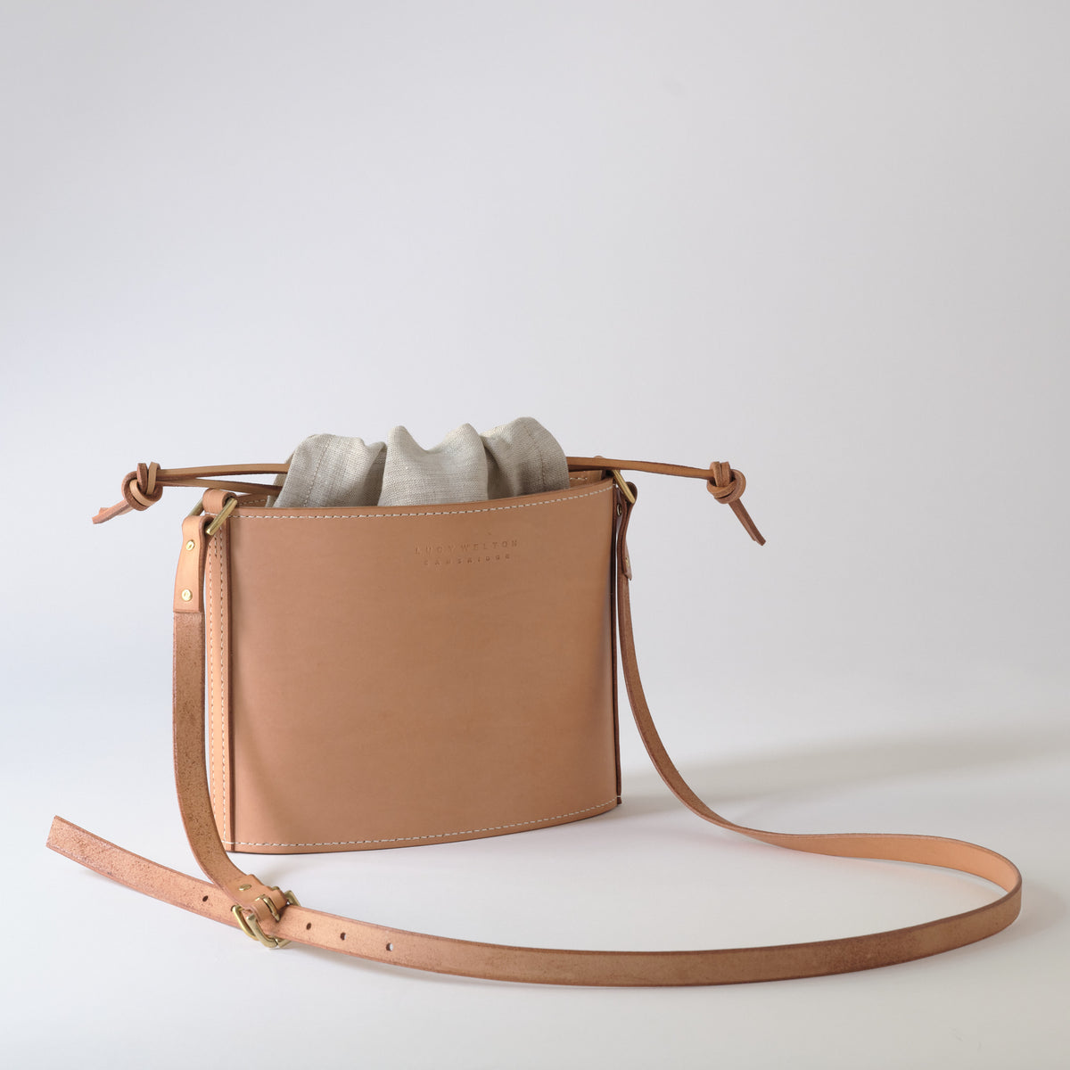 A light tan leather crossbody bag with a drawstring top and a long adjustable strap, shown on a clean light gray studio background.