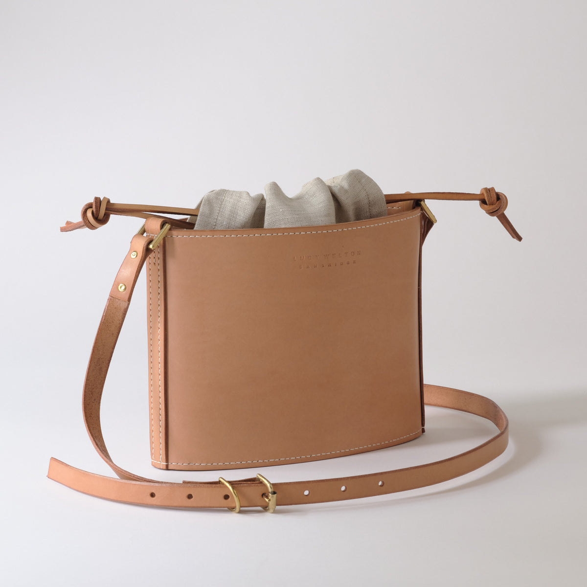 A tan leather crossbody bag with a drawstring top and a matching adjustable shoulder strap, shown on a clean light gray background. A light gray fabric dust bag is draped over the top of the bag.