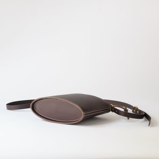A ldark leather crossbody bag with an oval body, a matching leather strap, and a small leather detail on the side, photographed on a light gray studio background.