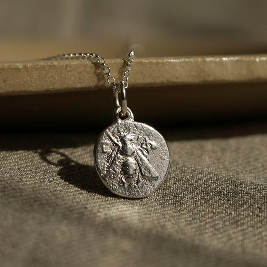 A silver coin pendant necklace featuring an embossed bee design, resting on a textured beige fabric surface with a ceramic dish rim in the background.