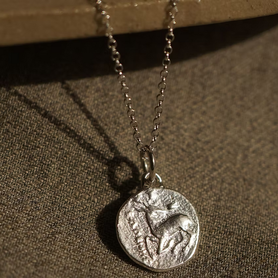 solid silver with a debossed picture of a stag in the middle on a silver chain necklace hanging on a ceramic plate on top of a hessian fabric
