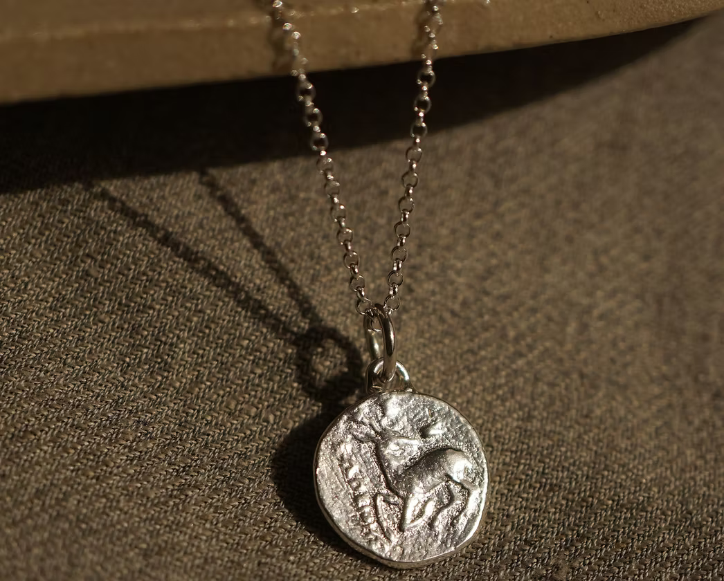 solid silver with a debossed picture of a stag in the middle on a silver chain necklace hanging on a ceramic plate on top of a hessian fabric
