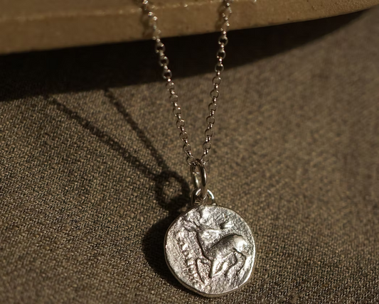 solid silver with a debossed picture of a stag in the middle on a silver chain necklace hanging on a ceramic plate on top of a hessian fabric