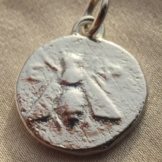 A close-up of a circular silver pendant or charm with a textured, antiqued finish of an illustrated bee and a small metal loop at the top, resting on a beige fabric background.