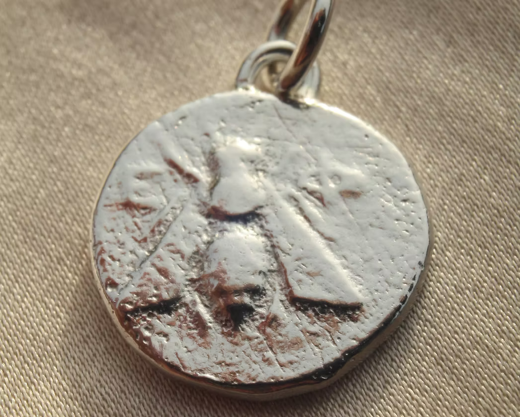 A close-up of a circular silver pendant or charm with a textured, antiqued finish of an illustrated bee and a small metal loop at the top, resting on a beige fabric background.