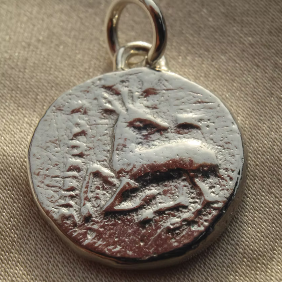 A close-up of a circular silver pendant or charm with a textured, antiqued finish of an illustrated stag and a small metal loop at the top, resting on a beige fabric background.