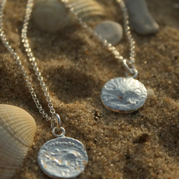 Two silver coin-style pendant necklaces on delicate chains, displayed on sand with seashells nearby.