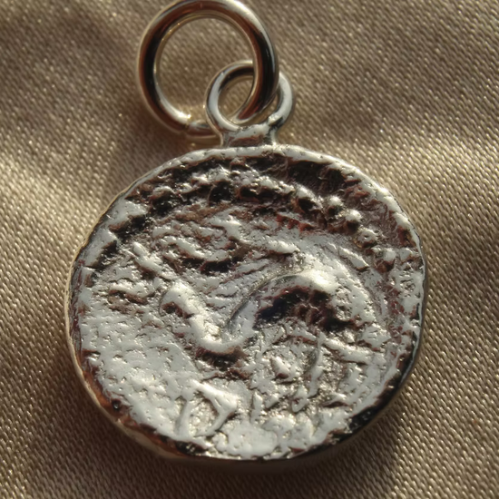A silver-toned ancient coin pendant charm with a textured, weathered finish and a small hanging loop, resting on a beige fabric background.