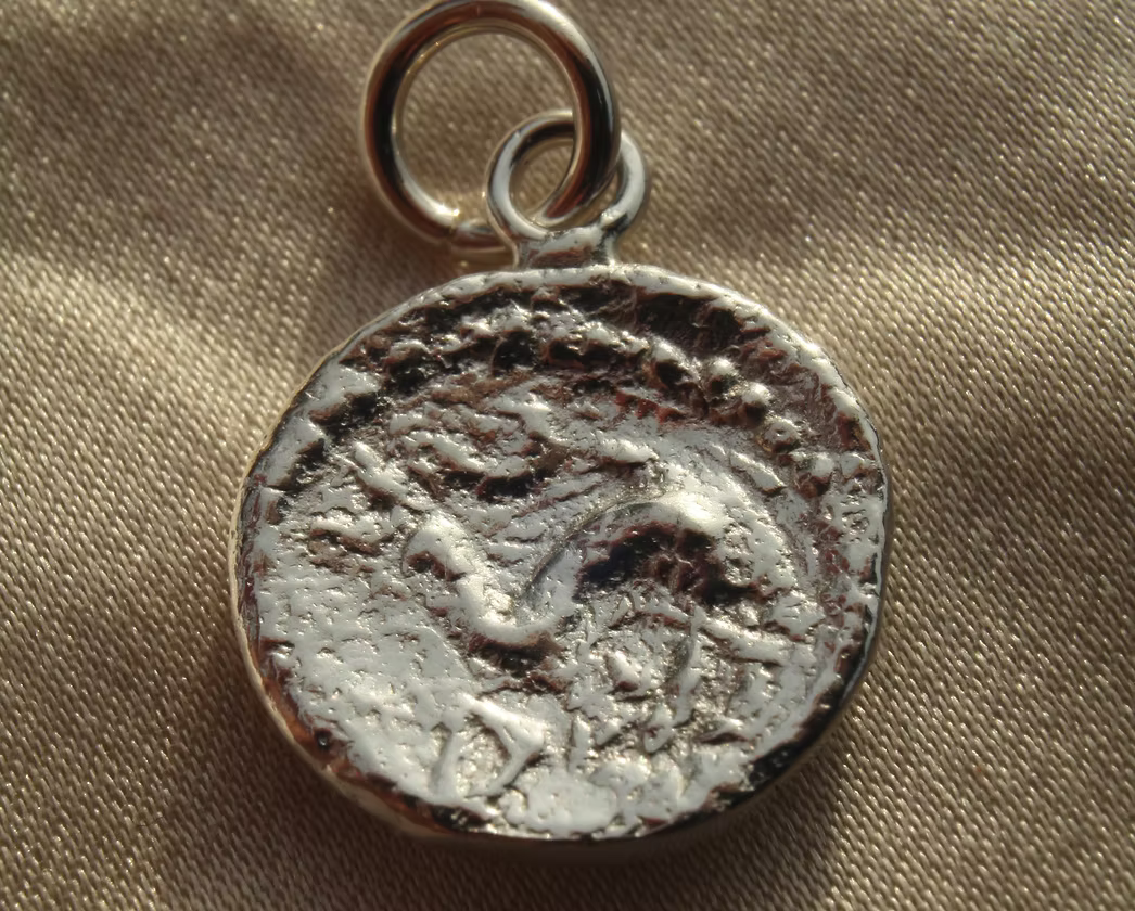 A silver-toned ancient coin pendant charm with a textured, weathered finish and a small hanging loop, resting on a beige fabric background.