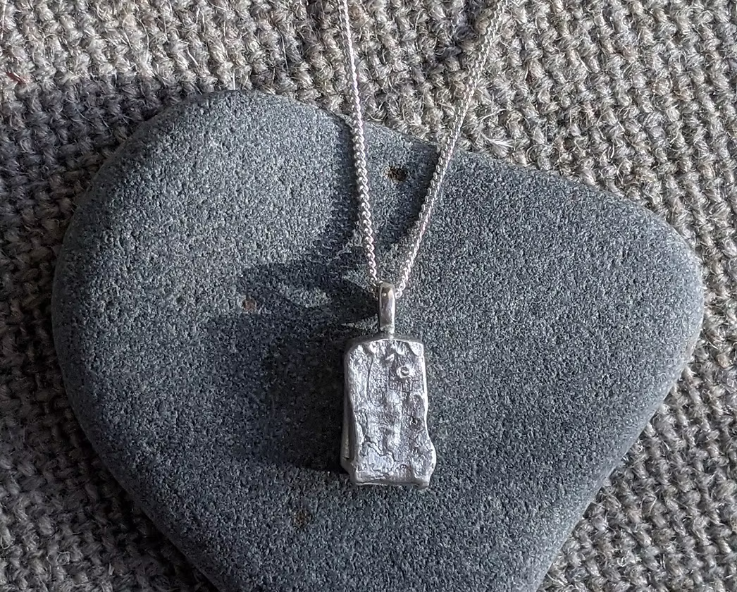 A silver pendant necklace with a textured, irregular rectangular pendant resting on a smooth grey stone, set against a woven beige fabric background.