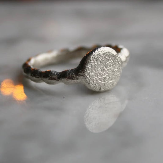 A silver ring with a textured surface and a rounded profile, displayed on a reflective surface with a blurred background.