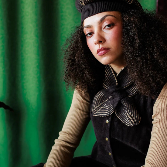 A model with curly dark hair is seated against a textured green backdrop, wearing a black beret, a beige ribbed long-sleeve top, a black corduroy vest with gold buttons, and a black bow tie with a beige feather pattern.