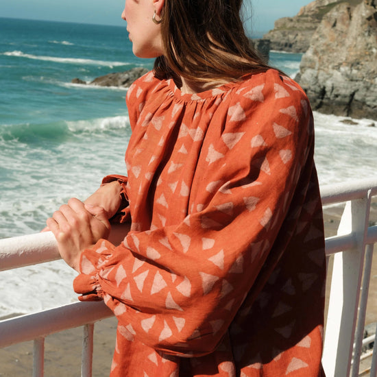 Woman in an orange dress with a geometric pattern standing by a railing overlooking the ocean.