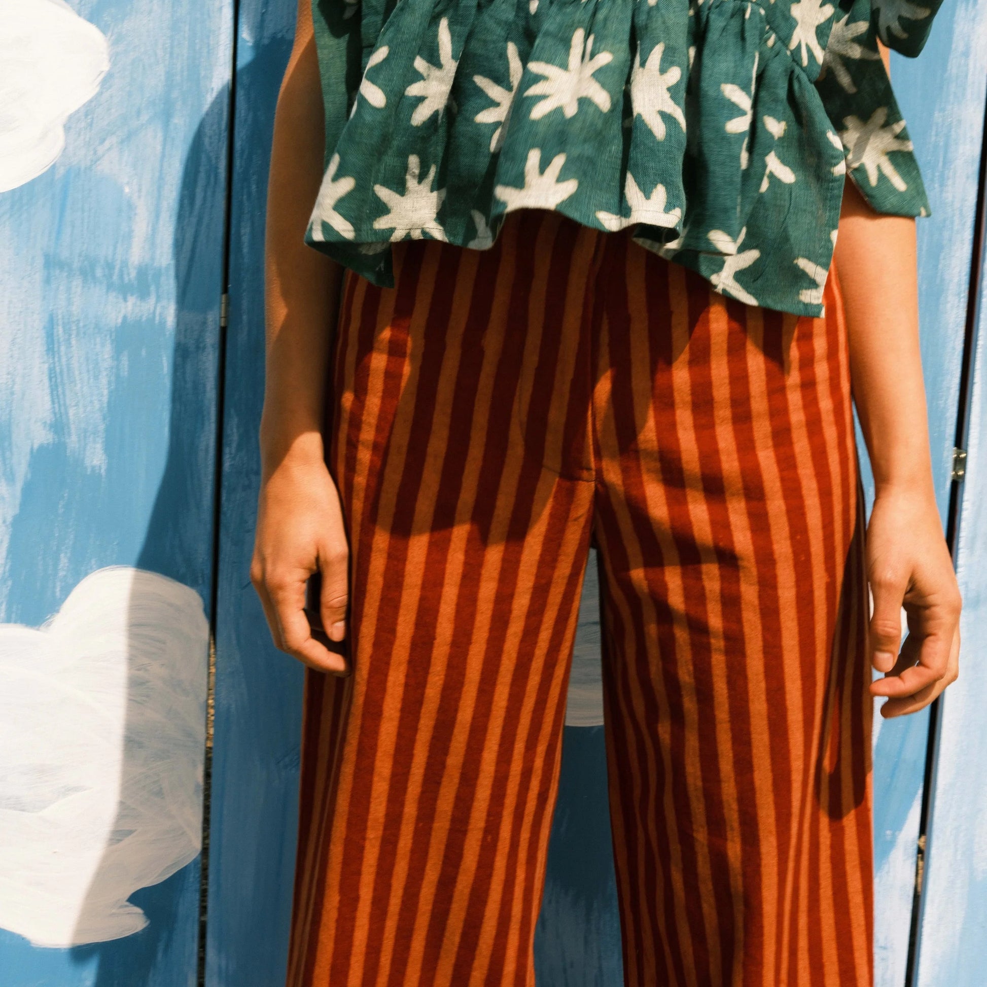 Person wearing a green and white floral top with ruffled layers and brown striped pants against a blue background.