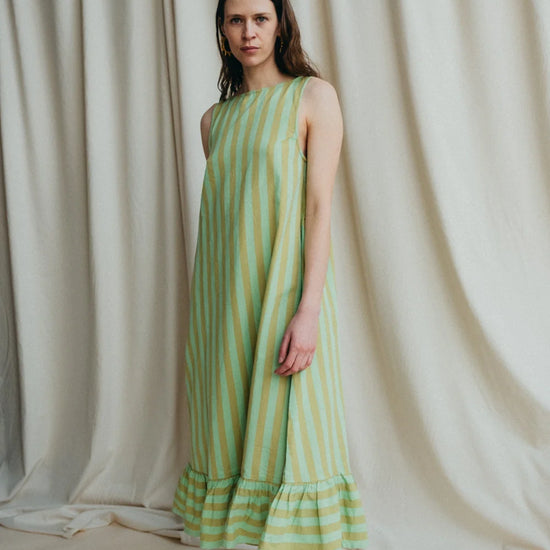 Model stands in green stripe sleeveless long dress with frill hem and bare feet against a a cream background 