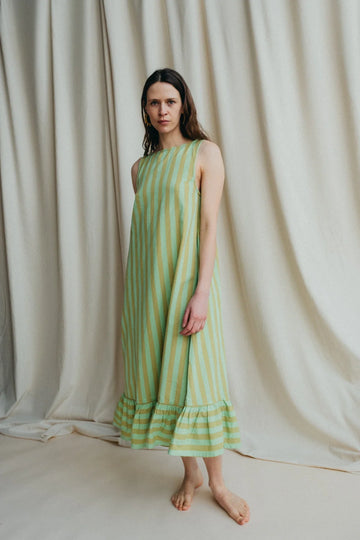 Model stands in green stripe sleeveless long dress with frill hem and bare feet against a a cream background 