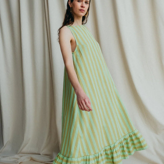 Model stands in green stripe sleeveless long dress with frill hem and bare feet against a a cream background 