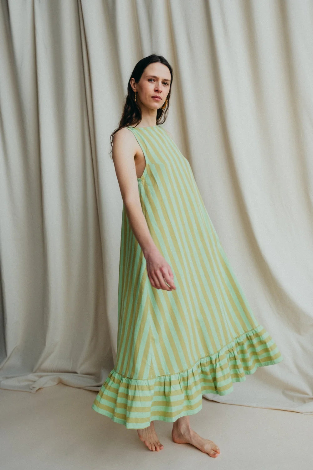 Model stands in green stripe sleeveless long dress with frill hem and bare feet against a a cream background 