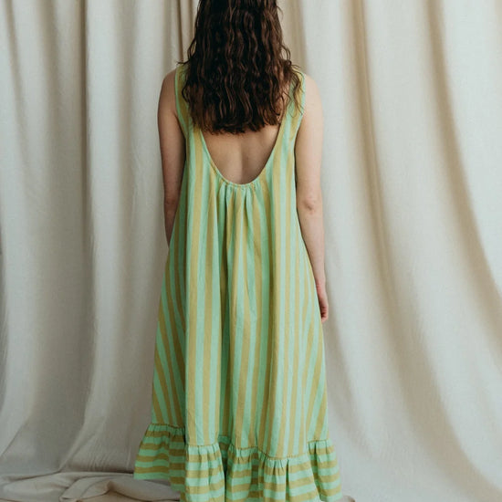 Model stands in green stripe sleeveless long dress to show the low back with frill hem and bare feet against a a cream background 