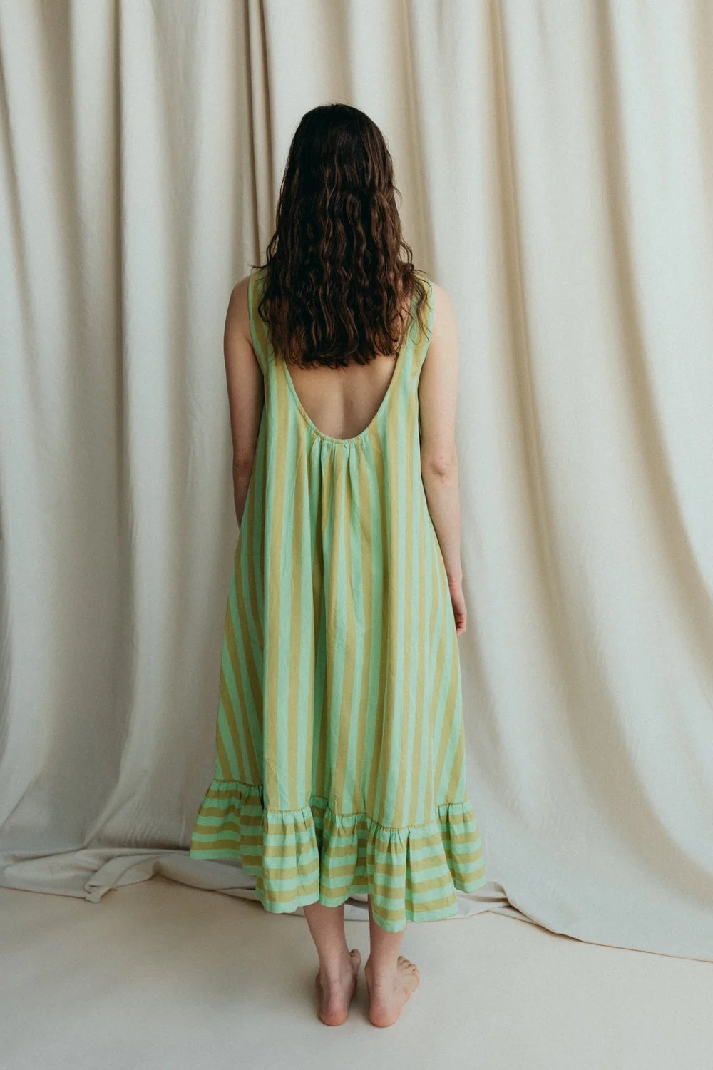 Model stands in green stripe sleeveless long dress to show the low back with frill hem and bare feet against a a cream background 