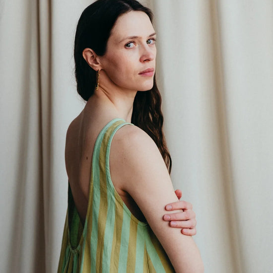 Model stands in green stripe sleeveless long dress to show the low back against a a cream background 