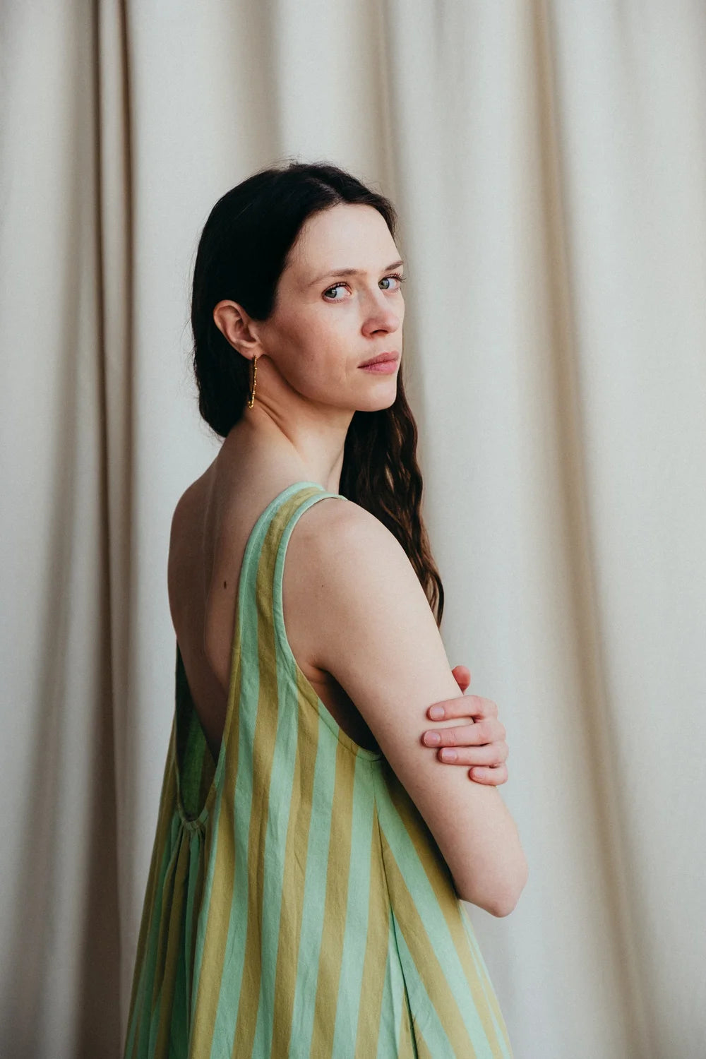 Model stands in green stripe sleeveless long dress to show the low back against a a cream background 