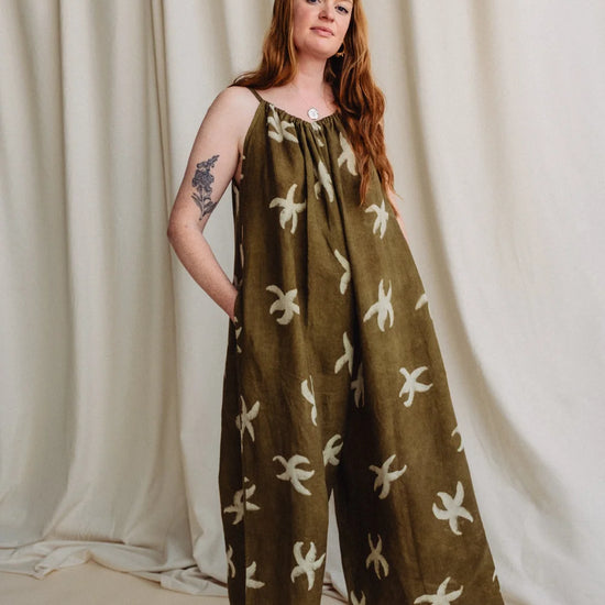 A woman modeling a wide-leg olive green jumpsuit with a cream abstract floral pattern, standing against a draped cream fabric backdrop.