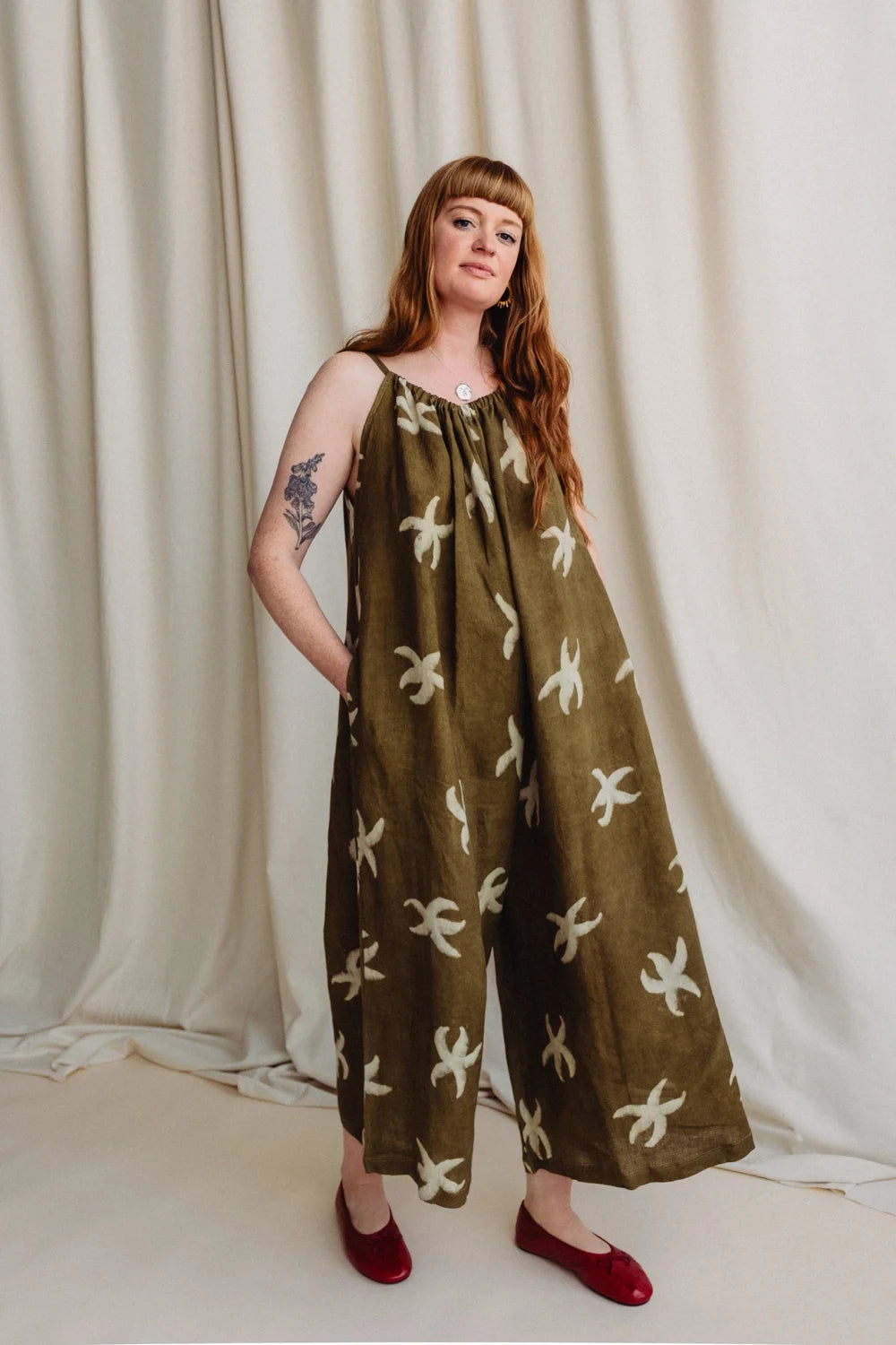 A woman modeling a wide-leg olive green jumpsuit with a cream abstract floral pattern, standing against a draped cream fabric backdrop.