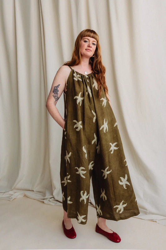 A woman modeling a wide-leg olive green jumpsuit with a cream abstract floral pattern, standing against a draped cream fabric backdrop.