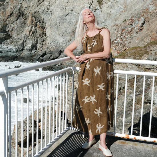 A woman with long platinum blonde hair stands on a coastal walkway wearing a brown sleeveless maxi dress with a white abstract pattern. The background features a large rocky cliff and the ocean.