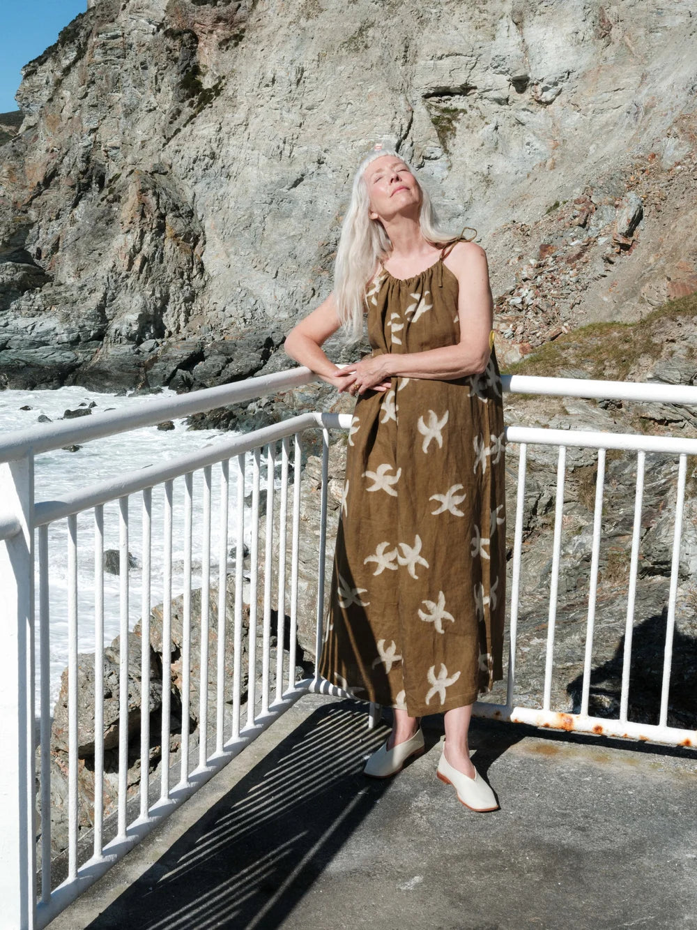A woman with long platinum blonde hair stands on a coastal walkway wearing a brown sleeveless maxi dress with a white abstract pattern. The background features a large rocky cliff and the ocean.