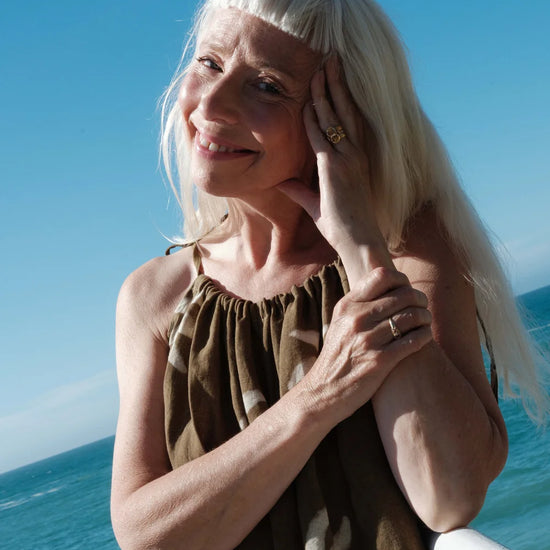 A smiling woman with long platinum blonde hair and a straight fringe, wearing an olive green strappy jumpsuit with a white abstract print, posing outdoors with the ocean and blue sky in the background.