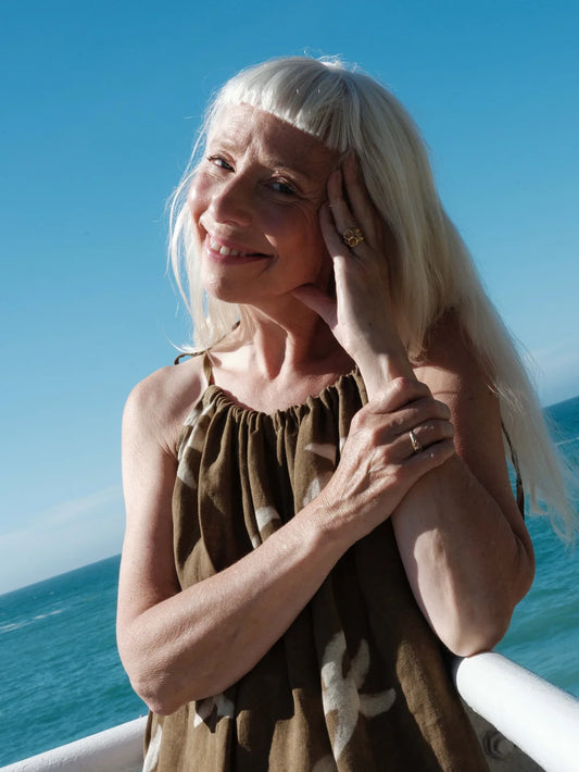 A smiling woman with long platinum blonde hair and a straight fringe, wearing an olive green strappy jumpsuit with a white abstract print, posing outdoors with the ocean and blue sky in the background.
