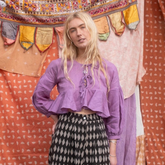 A woman standing in front of a colorful backdrop wearing a cropped purple top with balloon sleeves and a front tie detail, paired with black and white patterned pants.