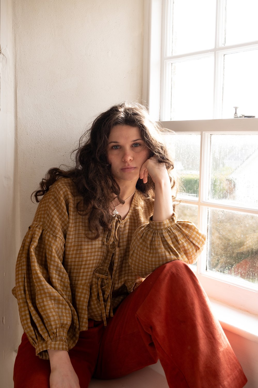 woman wearing flaxen linen top and red trousers sitting on window sill