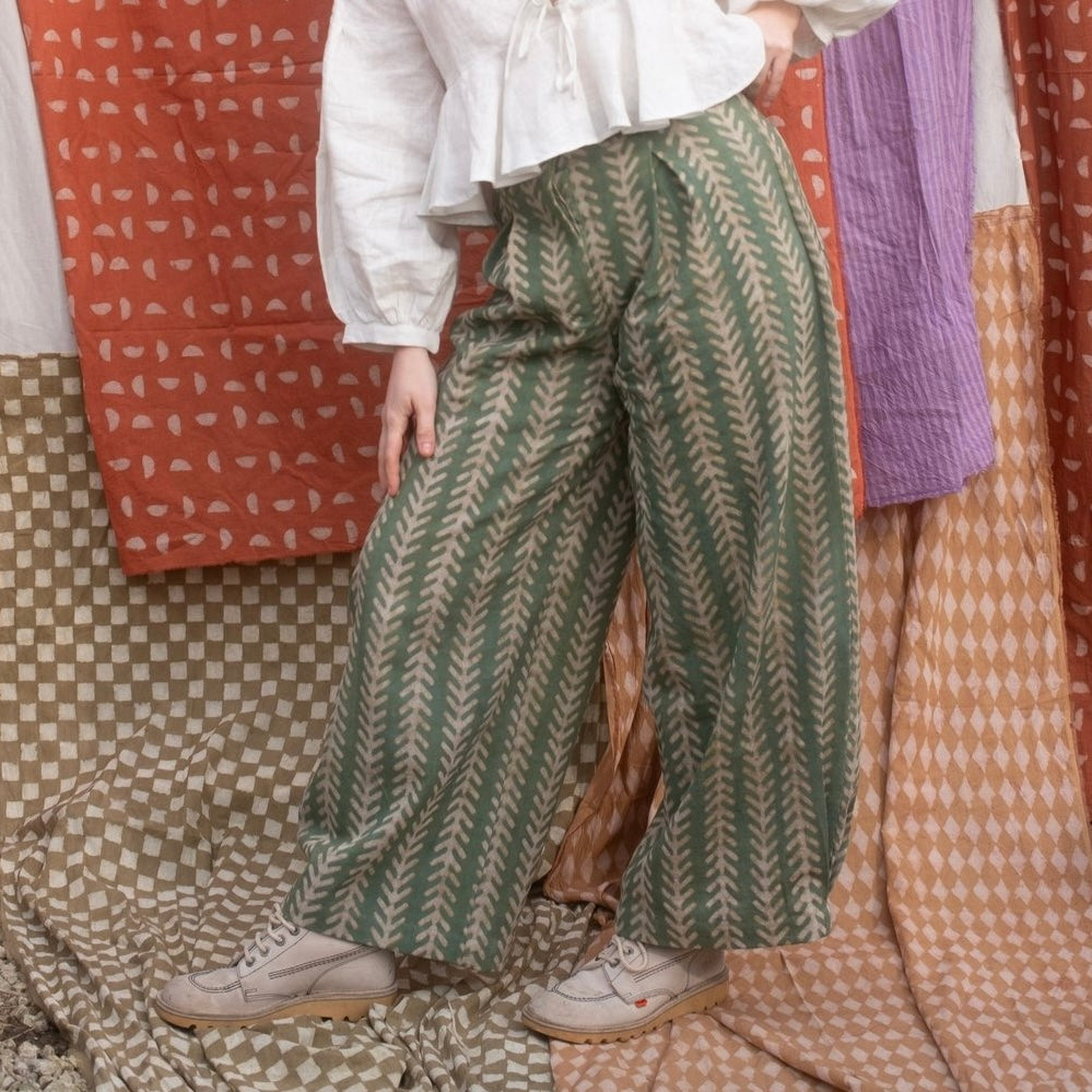 A person standing in front of a colorful backdrop wearing wide-leg green jeans with a geometric aloe print, a white shirt, and light-colored shoes.