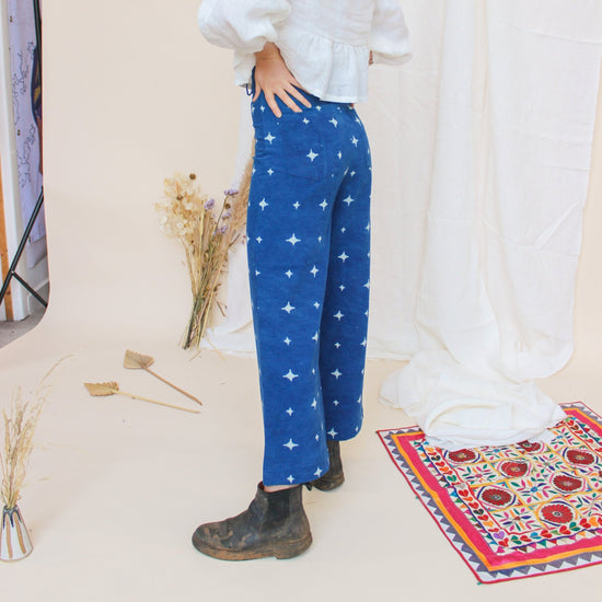 A person standing in a studio setting, wearing high-waisted indigo jeans with a star print, paired with a white top and black boots, in front of a white background with floral props.