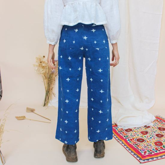 A person standing in a studio setting, wearing high-waisted indigo jeans with a star print, paired with a white top and black boots, in front of a white background with floral props.