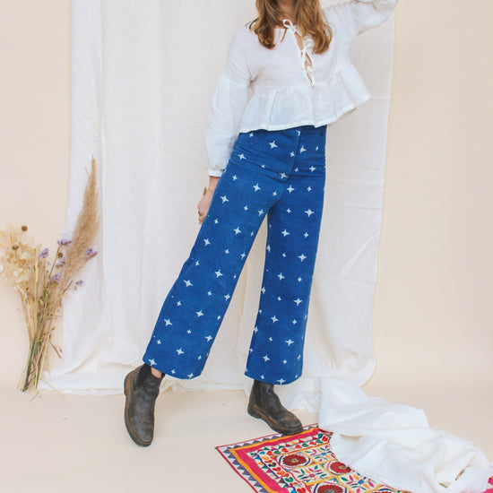 A person standing in a studio setting, wearing high-waisted indigo jeans with a star print, paired with a white top and black boots, in front of a white background with floral props.