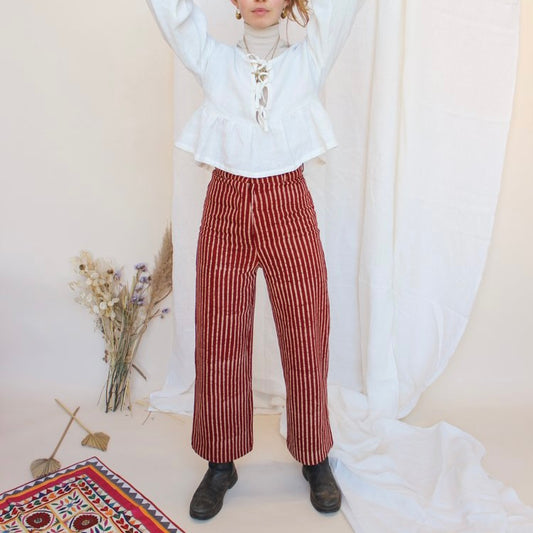 A person standing in front of a white fabric backdrop wearing high-waisted, red and white striped jeans with a white top and black boots. There is a small floral arrangement and a decorative, multicolored textile in the foreground.