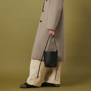 Woman wearing a long gray coat holding a black handbag against a green background