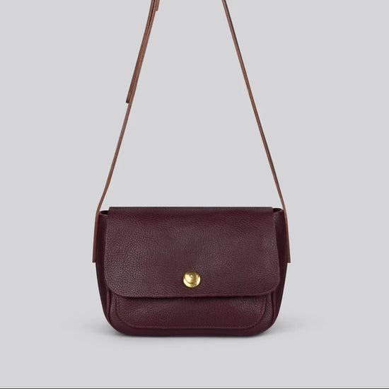 A small dark burgundy leather crossbody bag with a gold snap button and a thin shoulder strap, shown against a solid grey background.