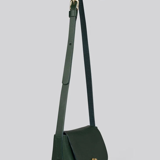 A dark green leather crossbody bag with a long adjustable strap and a small gold-tone turn-lock clasp, shown against a solid light gray background.
