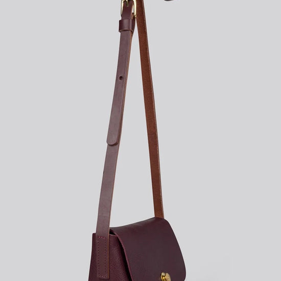 A dark burgundy leather crossbody bag with a matching adjustable shoulder strap and a small gold logo clasp on the front flap, shown against a solid grey background.